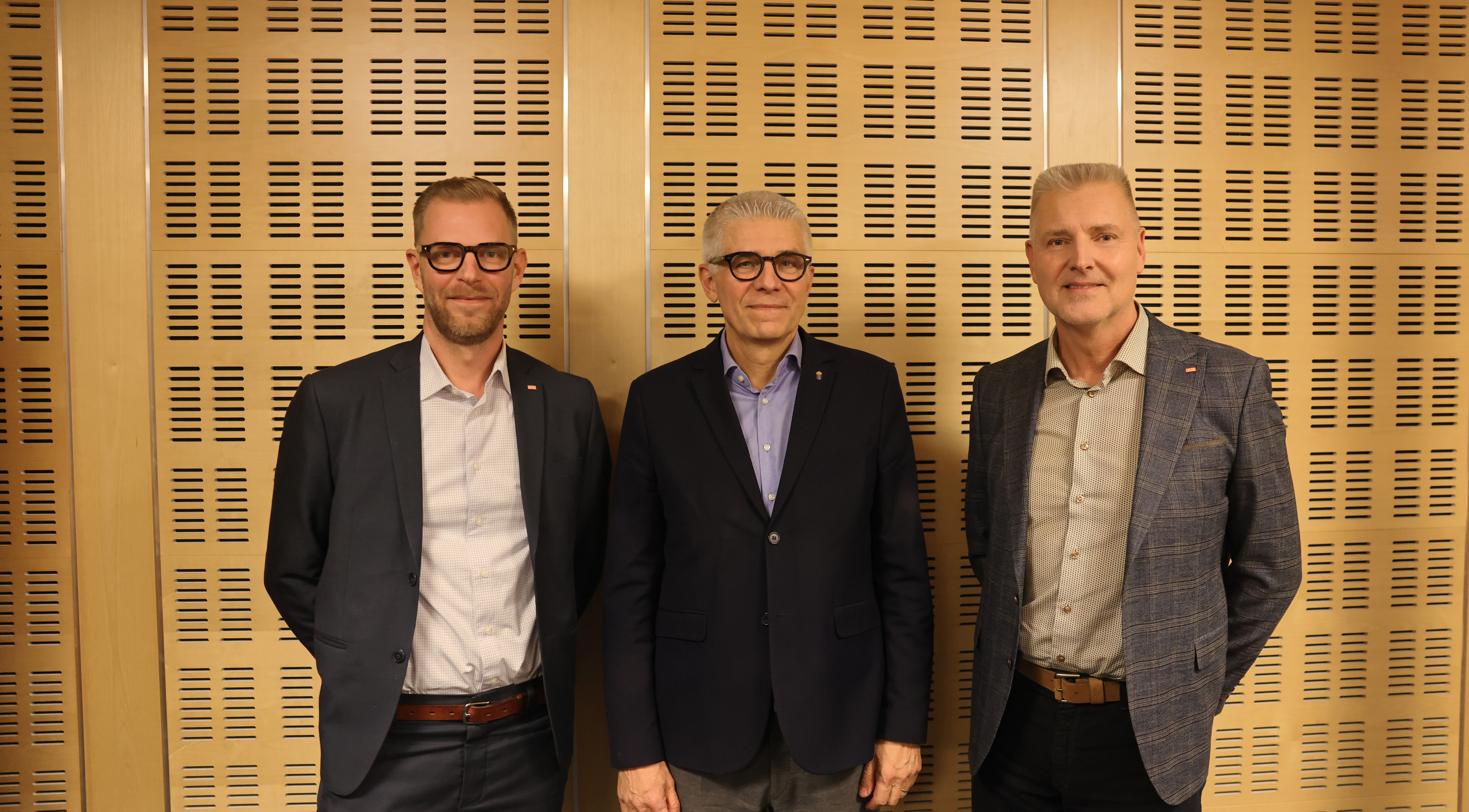 Tre män står bredvid varandra framför en vägg.