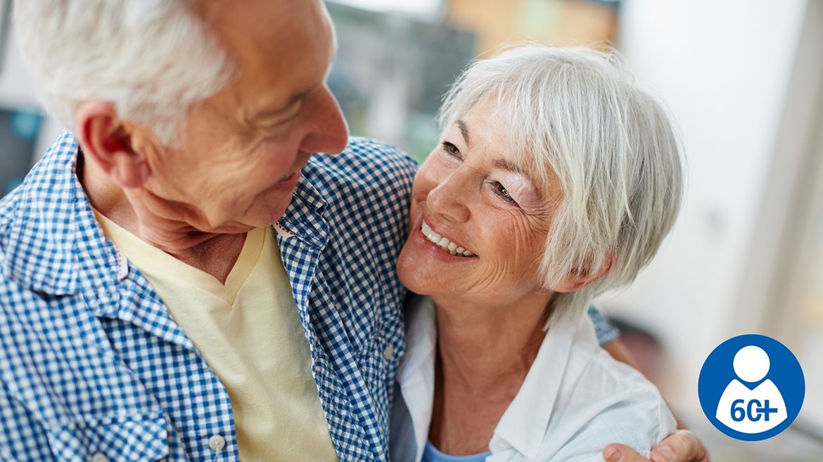 Seniorboende 60+. En äldre man håller sin arm om en äldre kvinna. De ler mot varandra.