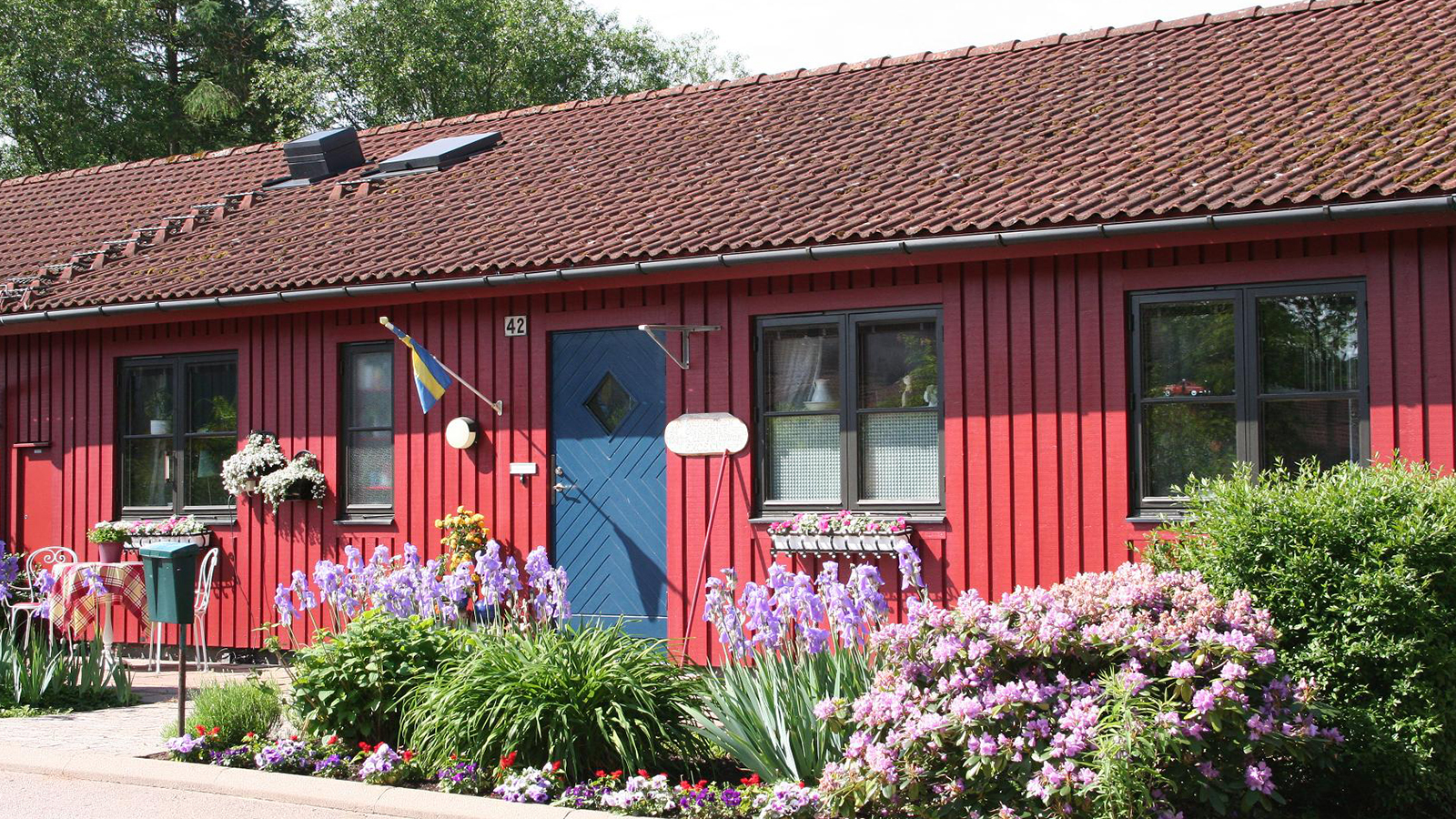 Frennarp. Sommarbild av ett rött enplansradhus med blå ytterdörr och prunkande rabatter.
